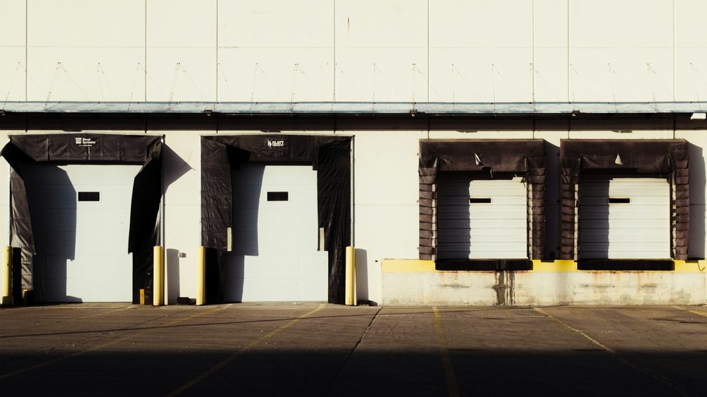 A row of closed loading dock doors along a warehouse wall in low sun, the yard in front of them empty.